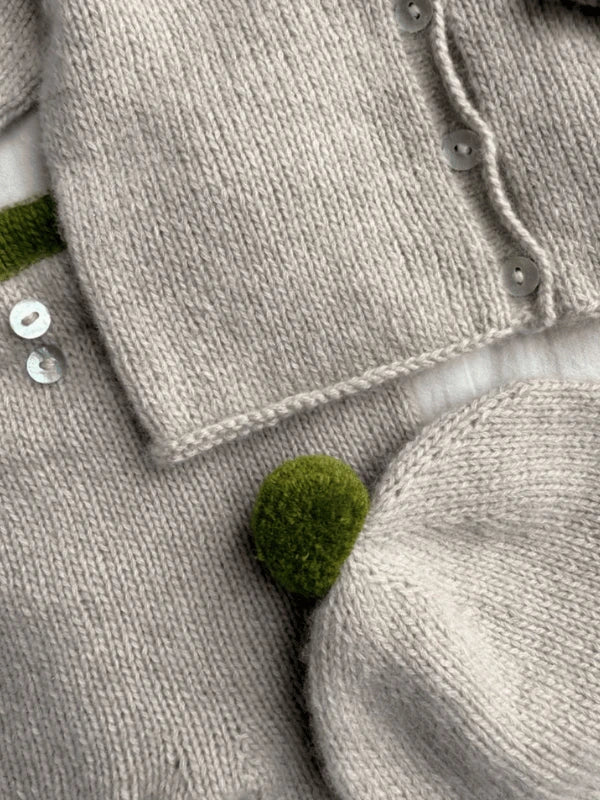 Close-up of soft, light gray knit fabric with white buttons and a green pom-pom from the Petiteknit Cleo Baby Set neuleohje Suomi, highlighting cozy vauvan neuleet and a simple, modern design.