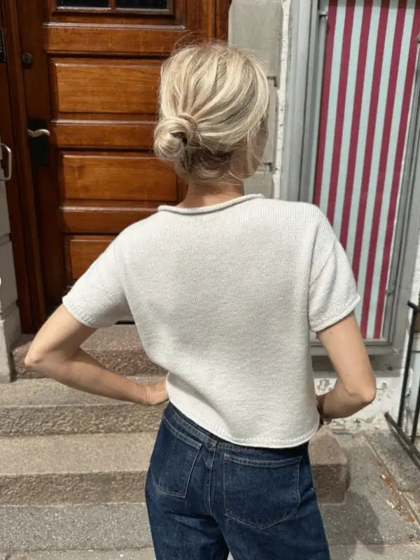 A woman with blonde hair in a low bun wears a short-sleeved white PetiteKnit Cloud tee neuleohje and dark blue jeans, standing with one hand on her hip, facing away from the camera by a wooden door and striped window.