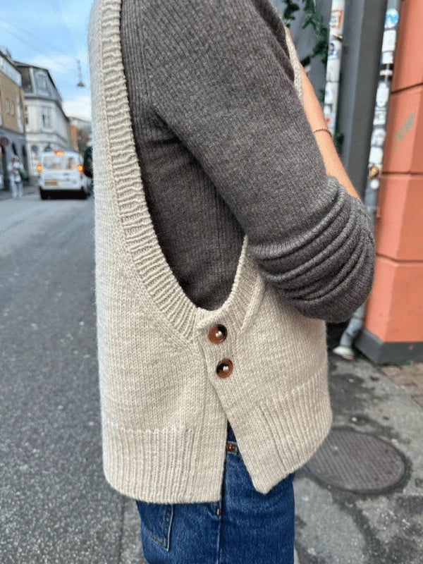 A person in a gray long-sleeve top, blue jeans, and the PetiteKnit - Lulu Slipover neuleliivi ohje with two brown side buttons stands on a city sidewalk near a peach-colored building.