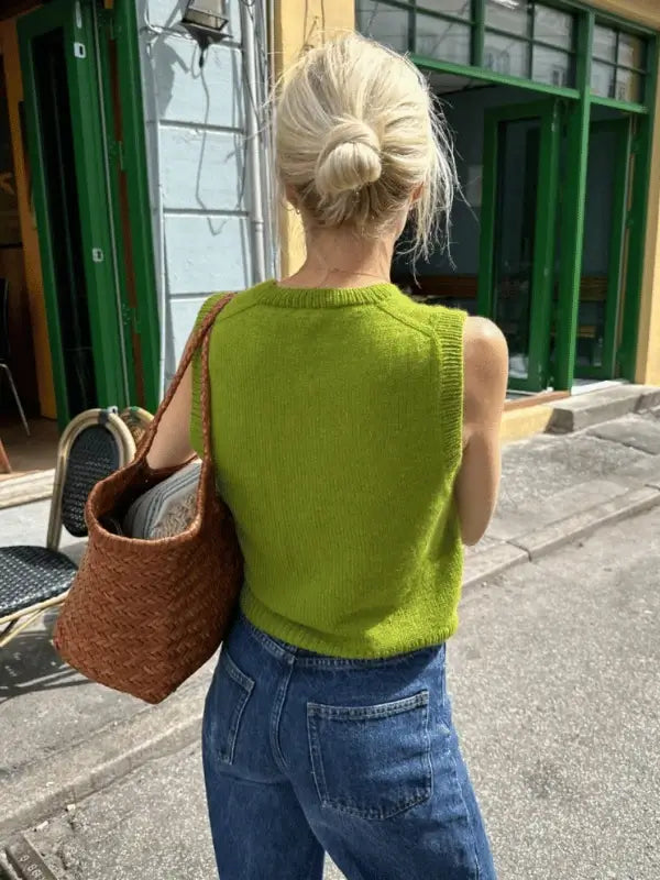 A woman with blond hair in a bun, wearing a sleeveless green PetiteKnit Maggie Vest neuleohje and blue jeans, carries a woven brown shoulder bag while walking past green-framed windows and outdoor chairs on a sunny street.