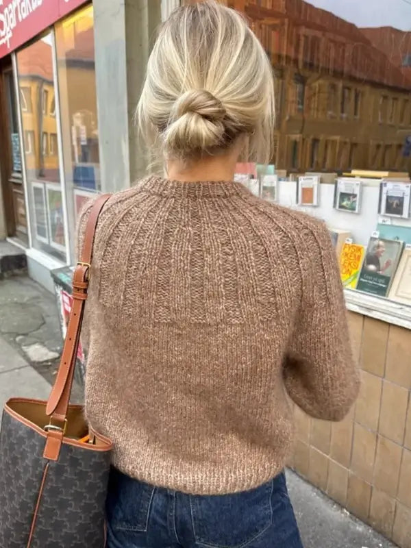 A woman with blonde hair in a low bun, wearing the PetiteKnit Sunday Sweater neuleohje, Suomi in brown and blue jeans, stands on a city sidewalk with a patterned shoulder bag, facing a shop window displaying cards.