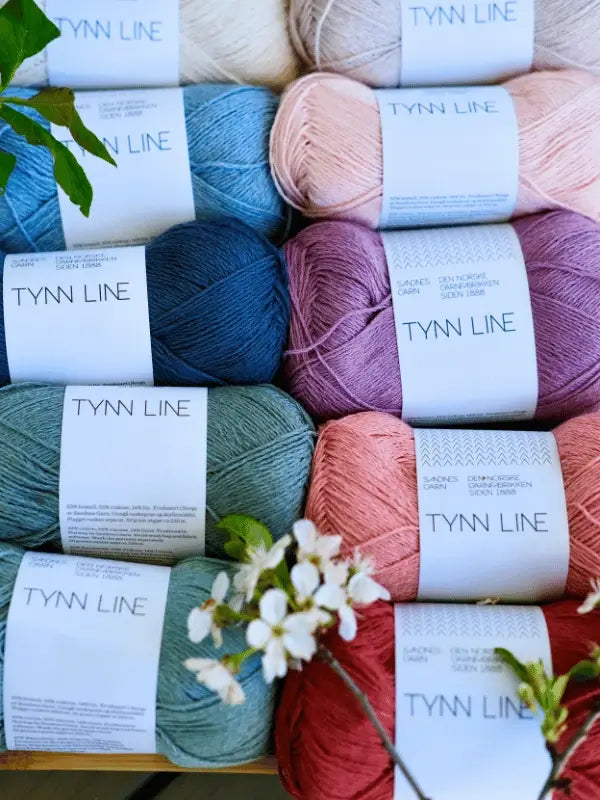 Several skeins of Sandnes Garn Tynn Line yarn in soft pastel colors are arranged in rows with green leaves and white flowers, making them perfect for your next summer knit project.