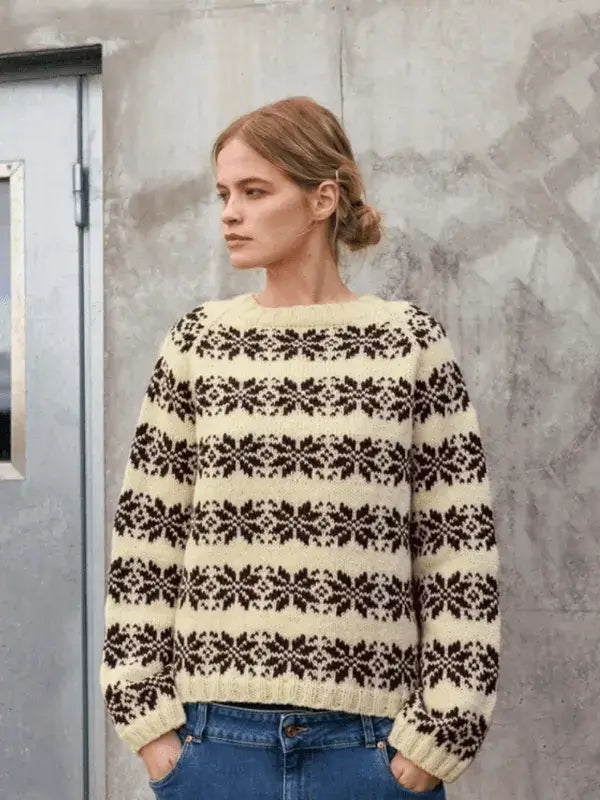 A woman wearing a cream-colored Sandnes Garn Alvar – neulesetti Atlas featuring a brown, repeating floral pattern across the chest, sleeves, and shoulders.