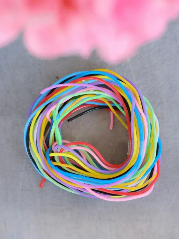 A coil of colorful licorice candy strings, resembling Sandnes Garn Silmukkakaapelit, is arranged in a loose circle on a gray surface with a blurred pink object at the top.