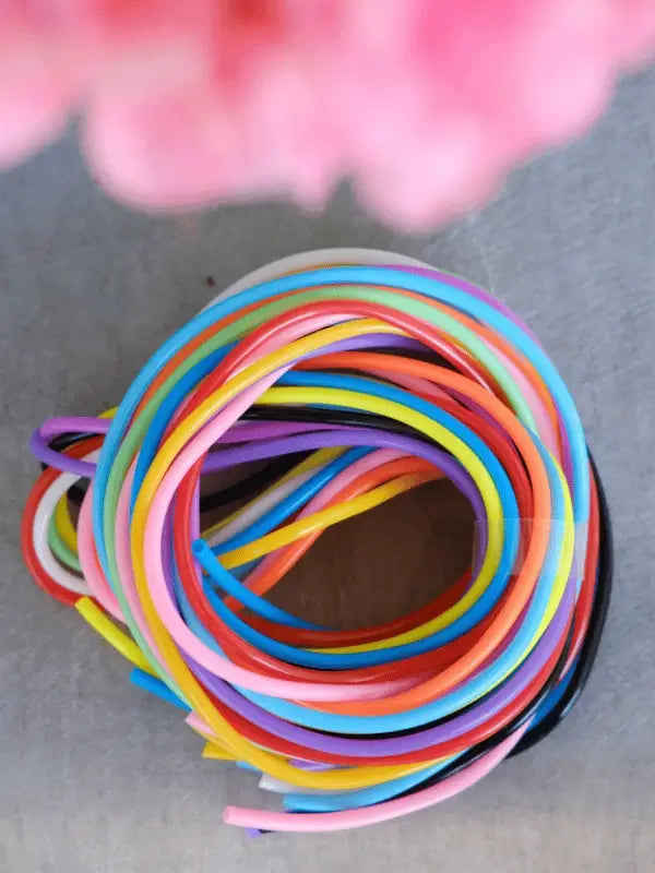 A bundle of Sandnes Garn Silmukkakaapelit, colorful plastic-coated wires for neulominen and as a handy neuleapu, is coiled on a gray surface viewed from above, with a pink blurred object at the image's top.