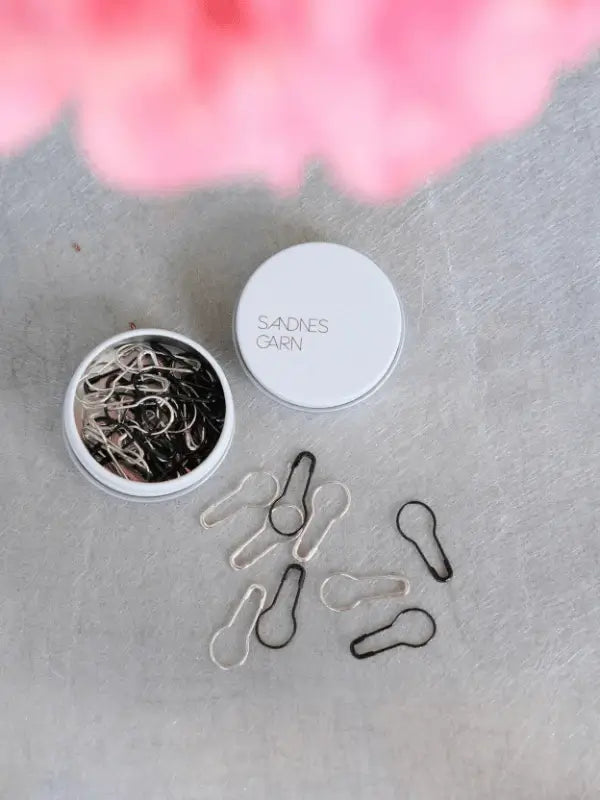 A round white tin labeled "Sandnes Garn Silmukkamerkit" holds black and silver bulb-shaped stitch markers, with several scattered on a gray surface. Pink blurred petals are visible at the top of the image.