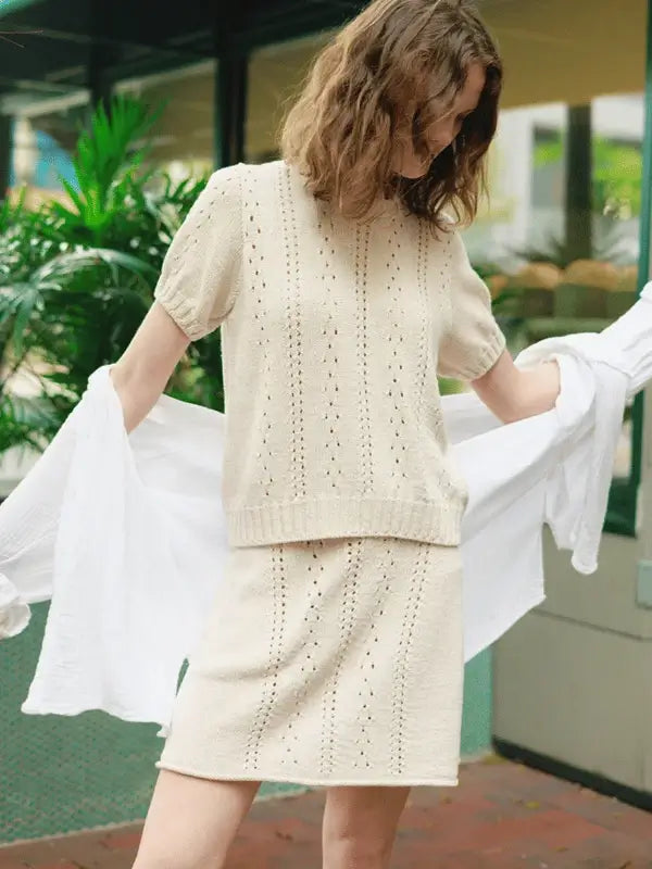 A woman wearing the Cornelia Tee & Skirt in cream-colored Sandnes Garn Line stands outside holding a white fabric. Green plants and a building with large windows are visible in the background.