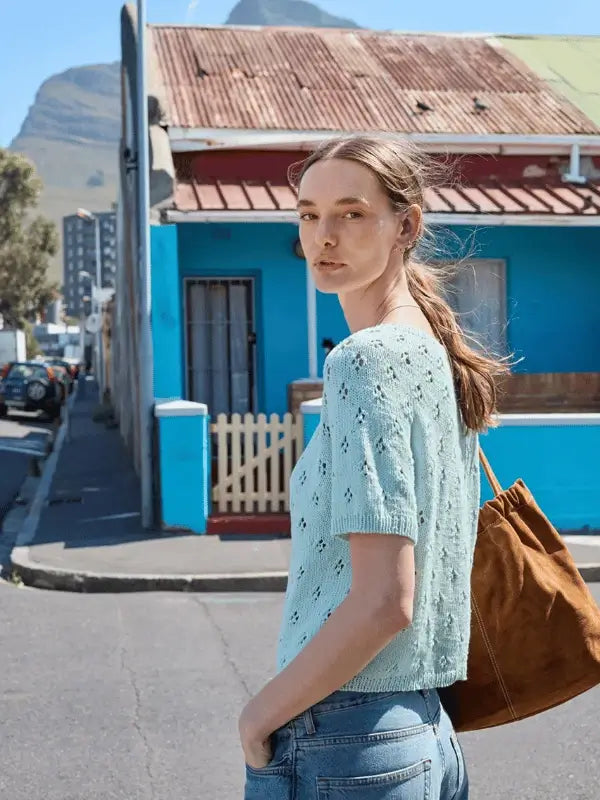 A woman with light brown hair in a low ponytail stands outdoors on a sunny day, wearing jeans and the Elodie Tee knitted from Sandnes Garn Line, holding a brown bag in front of a bright blue house.