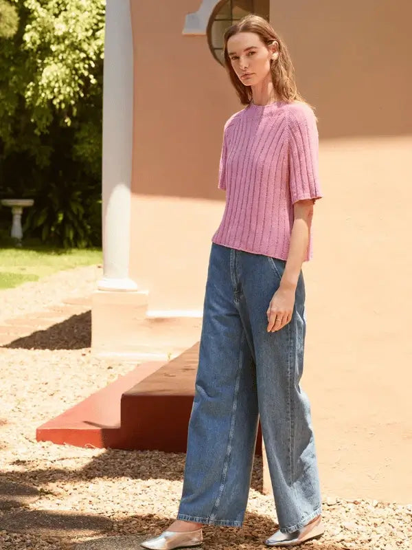 A woman stands outdoors in the pink Elva tee neuleohje ENG, knitted with Sandnes Garn Line, paired with wide-leg blue jeans and silver flats, beside a peach-colored building with sunlight and greenery behind her.