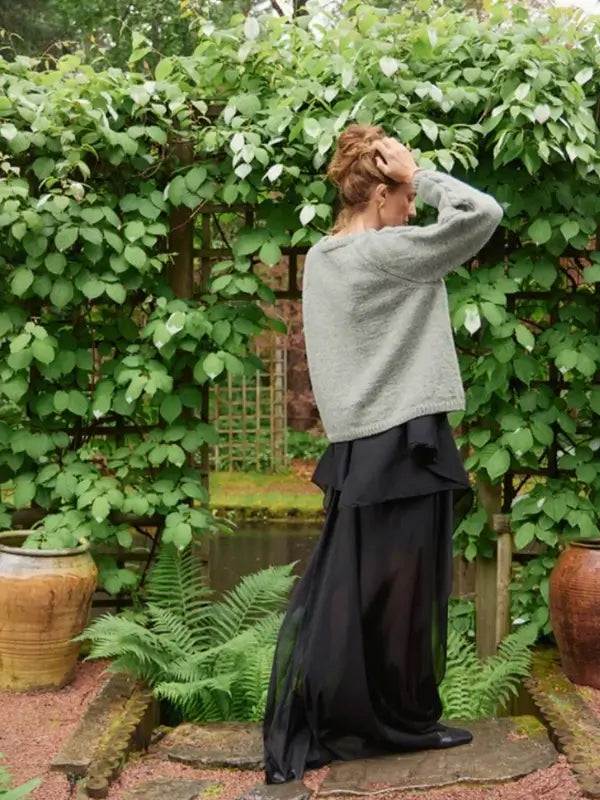 Wearing a cozy gray Laura Sweater made with the Sandnes Garn 2606-10 DIY Laura Sweater pattern (neuleohje suomeksi, PDF), a person stands outdoors facing sideways in a long black sheer skirt. With one hand on their head, they are surrounded by lush green plants and large clay pots.
