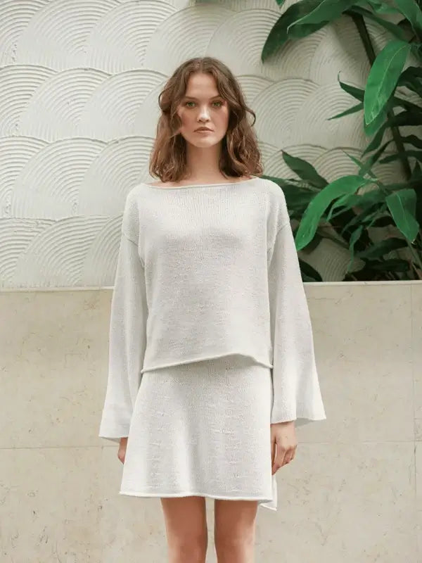 A woman stands indoors in the Milly Sweater & Skirt Neuleohje Suomi + Sandnes garn line, her wavy hair and calm expression framed by green plants and textured white walls for a serene käsityö-inspired look.