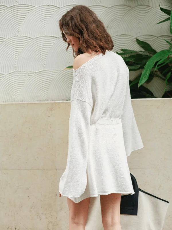 A woman with wavy brown hair wears the loose-fitting, white Milly Sweater from the Neuleohje Suomi + Sandnes Garn Line, styled off-shoulder with long sleeves. She stands sideways, looking down in front of a textured wall and green plants.