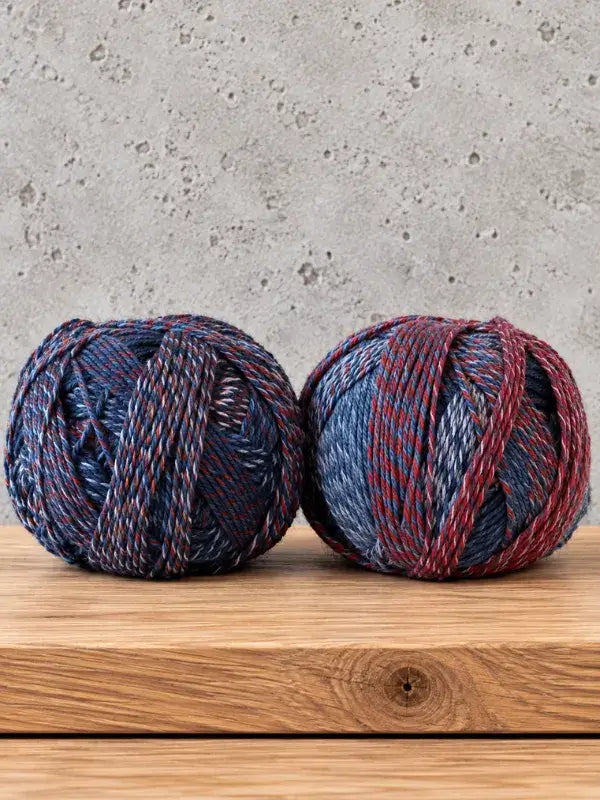 Two balls of Schoppel-Wolle Edition 3 yarn in a variegated blue, red, and off-white pattern resting on a wooden surface.