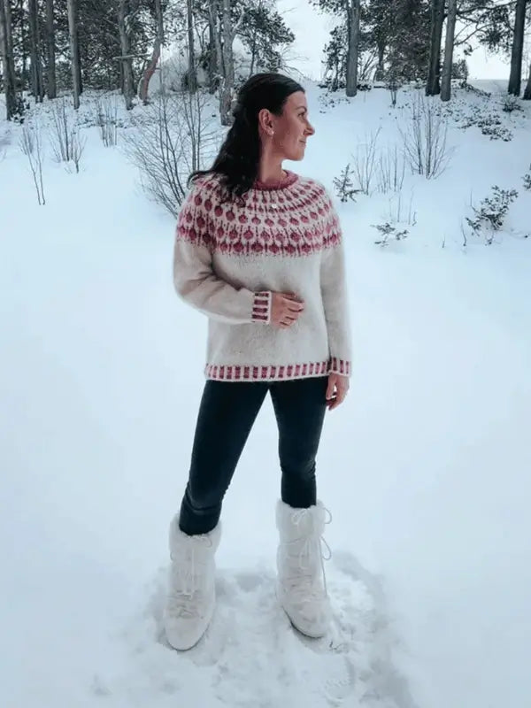 A woman stands in the snow wearing a cozy white Sisu Knitwear Vadelmaneule neuleohje Suomii PDF sweater with red patterns, black pants, and white winter boots. Snow-covered trees and bushes are in the background.