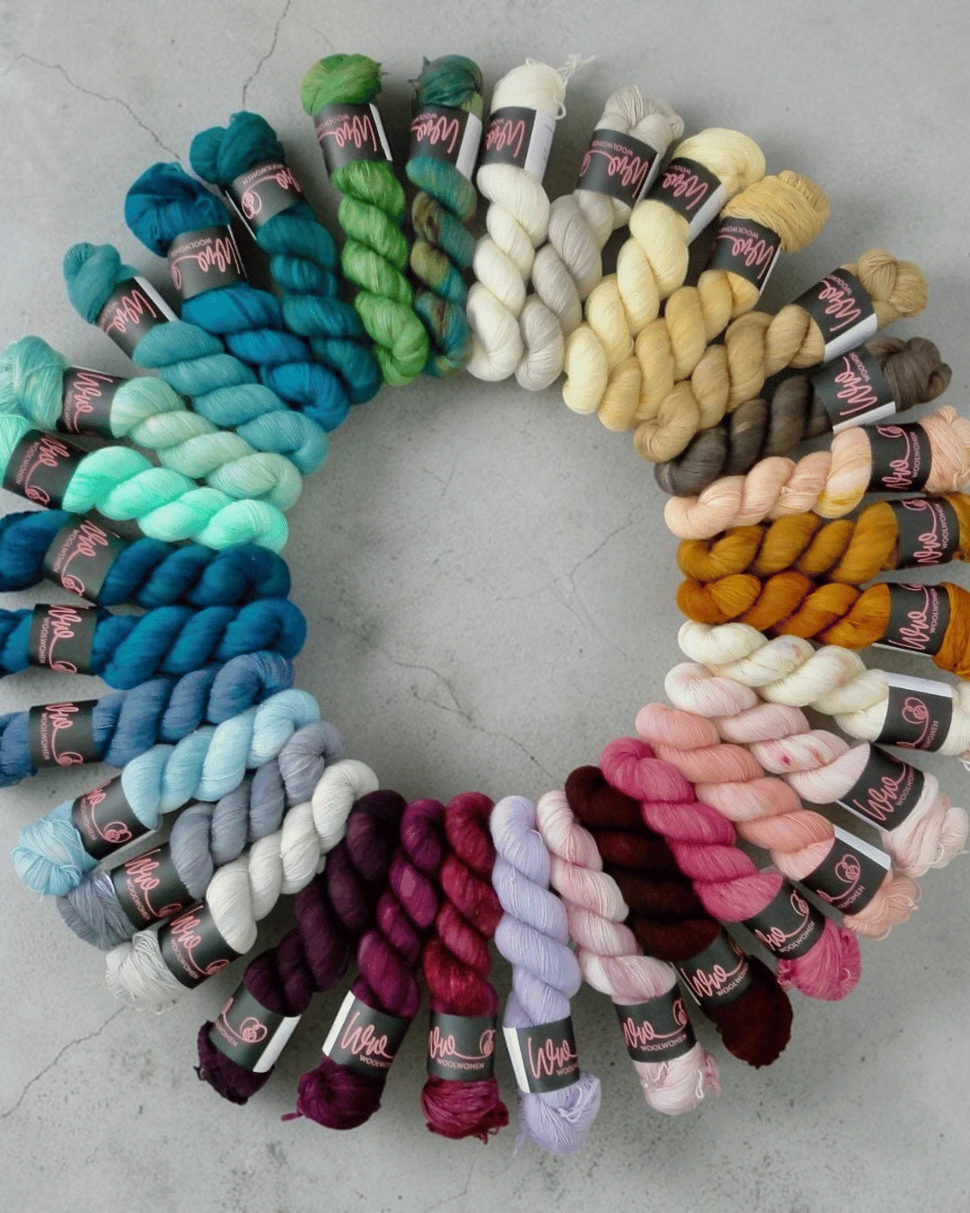 A circular gradient of WoolWomen Merino Single skeins, arranged from dark greens and blues to cream, mustard, browns, reds, pinks, and purples, displayed on a light gray surface.