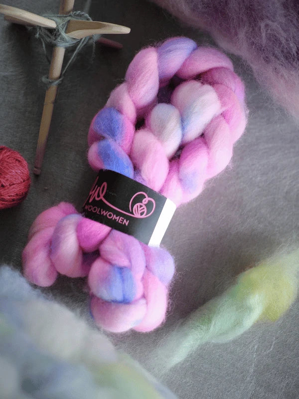 Two bundles of WoolWomen kehruukuitu Corriedale käsinvärjätty in pink, purple, and white rest on a grey surface with a wooden spindle and green wool, ideal for kehräämiseen projects.