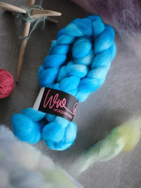 Two bundles of bright blue, chunky WoolWomen kehruukuitu Corriedale käsinvärjätty with black Wool Women labels lie on a gray surface, surrounded by pieces of soft green fiber and a white decorative star.