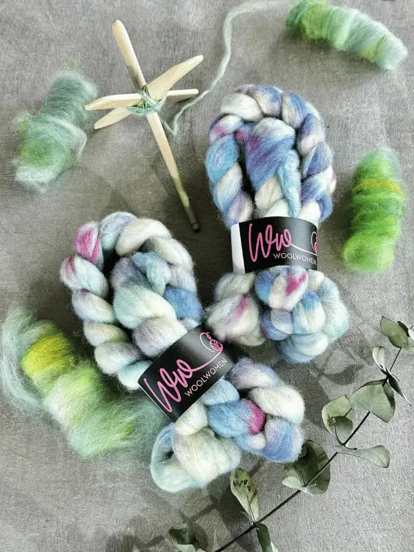 Two skeins of WoolWomen Kehruuvilla BFL Käsinvärjätyt wool roving in blue, white, and pink rest on a gray surface with green wool, a wooden spindle, and eucalyptus—ideal käsinvärjätty kehruukuitu kehräämiseen.