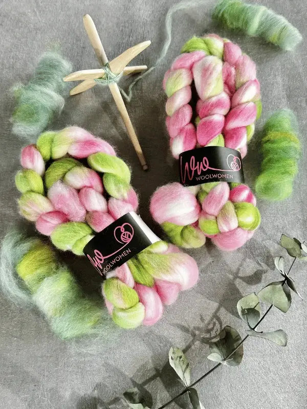 Two skeins of WoolWomen kehruukuitu Polwarth käsinvärjätty in pink, green, and white sit on a gray surface near fluffed wool, a wooden drop spindle for kehrääminen, and eucalyptus sprigs.