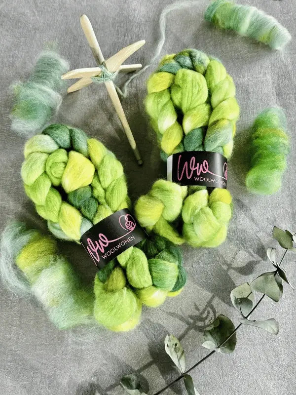 Two braids of WoolWomen kehruukuitu Polwarth käsinvärjätty in green and yellow, with black labels, rest on a gray surface amid loose wool, a wooden spinning spindle, and dried eucalyptus leaves.