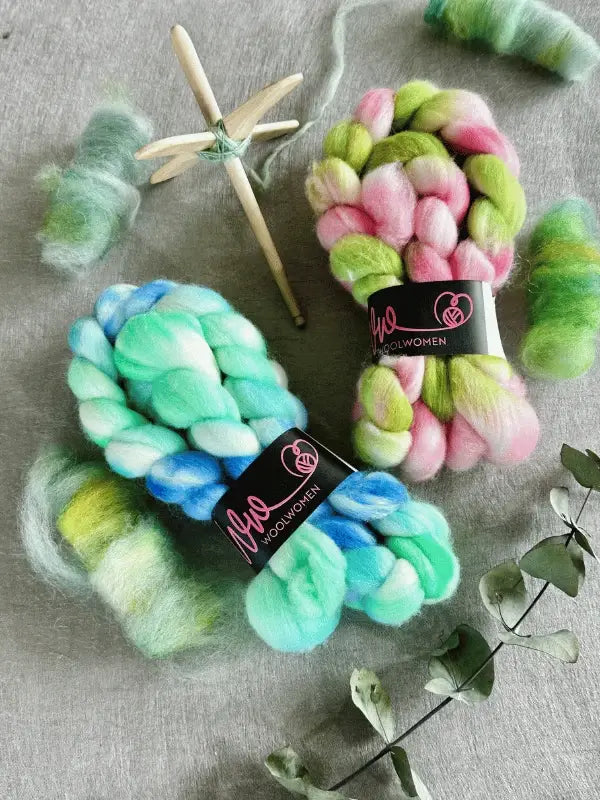 Two braided bundles of WoolWomen kehruukuitu Polwarth käsinvärjätty—one blue-green, one pink-green—are displayed on fabric with loose wool, a wooden drop spindle, and dried eucalyptus leaves nearby.