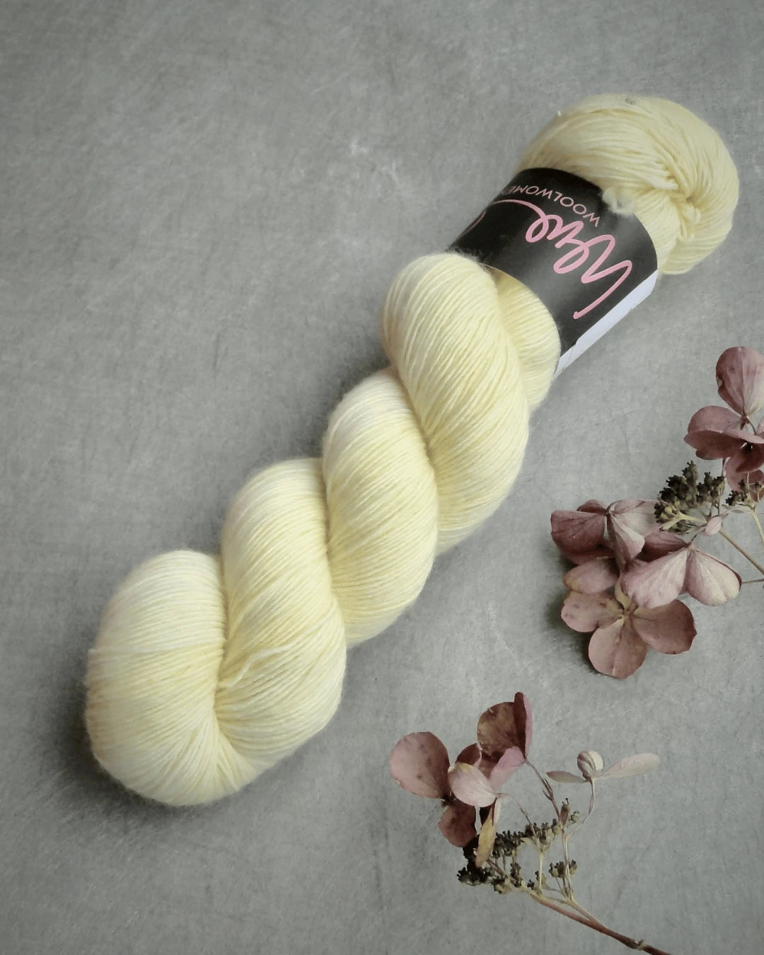 A skein of pale yellow WoolWomen Merino Single yarn with a black label rests on a gray surface beside dried, light pink hydrangea flowers.