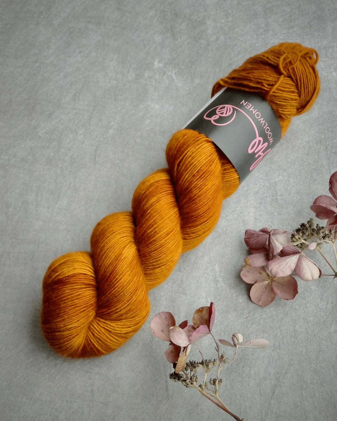 A skein of WoolWomen Merino Single käsinvärjätty merinovilla mustard yellow yarn rests on a gray surface next to dried pink and beige flowers. The skein features a black label with pink text and a logo.