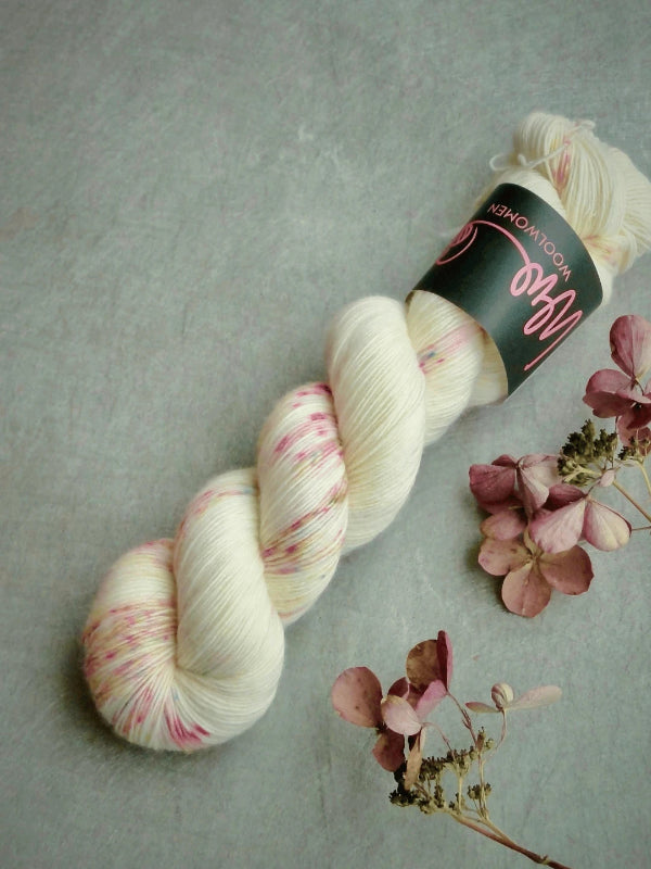 A skein of WoolWomen Merino Single, käsinvärjätty merinovilla in pale cream with pink speckles, is displayed on a gray surface beside dried pink and beige hydrangea flowers.