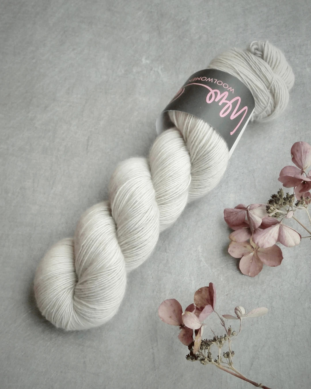 A skein of WoolWomen Merino Single, soft light gray yarn with a black and pink label, rests on a gray surface beside dried pink hydrangea flowers.