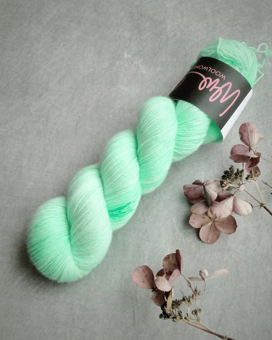 A skein of mint green WoolWomen Merino Single yarn lies on a light grey surface next to dried hydrangea flowers. The yarn features a black label with pink and white text.