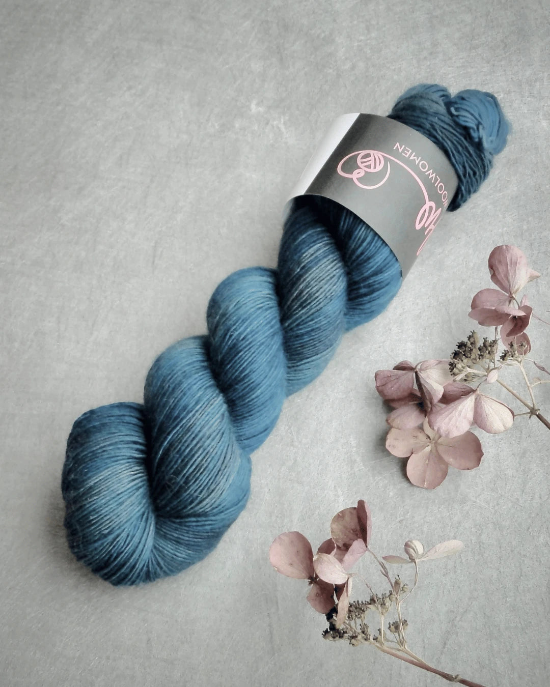 A skein of WoolWomen Merino Single, käsinvärjätty blue merinovilla yarn with a label, rests on a gray surface beside dried pink and beige flowers.