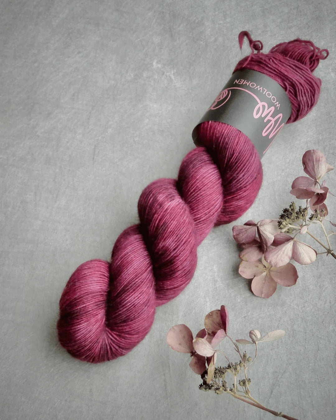 A skein of magenta WoolWomen Merino Single yarn lies on a gray surface beside light pink dried flowers, with its label wrapped around it.