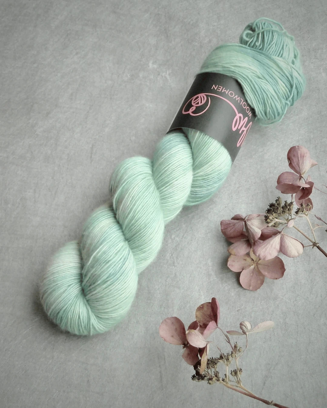 A skein of pale mint green WoolWomen Merino Single yarn with a black label rests on a gray surface next to dried pink hydrangea flowers.