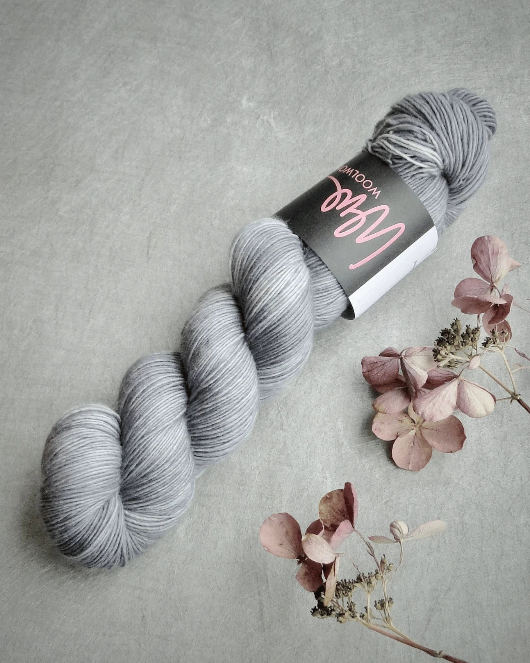 A skein of WoolWomen Merino Single yarn in light grey with a black and pink label rests on a grey surface beside delicate dried pink flowers.