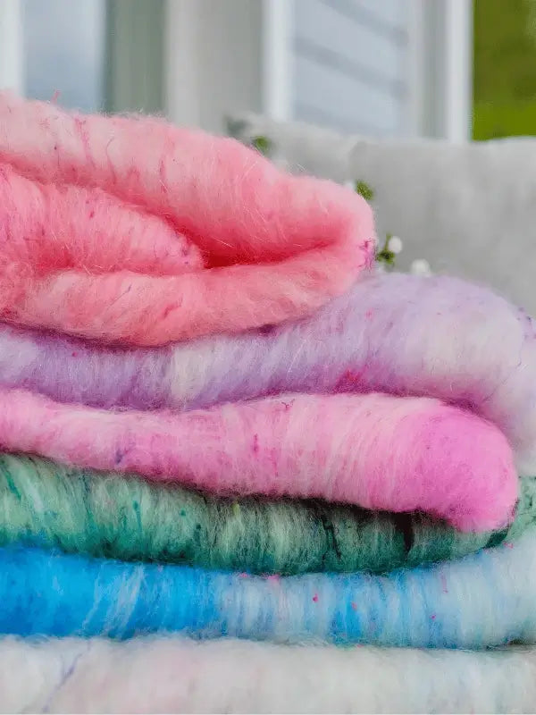 A close-up of a neatly stacked pile of WoolWomen Art Batt -Corriedale in pastel shades—pink, lilac, green, and blue—crafted from käsinvärjätty villa. The softly blurred backdrop reveals hints of a window and outdoor greenery.