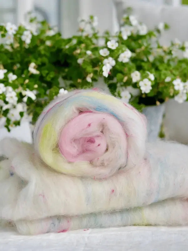 A roll of WoolWomen Art Batt -Corriedale, soft and multicolored, rests on folded wool with a potted plant of small white flowers softly blurred in the background.