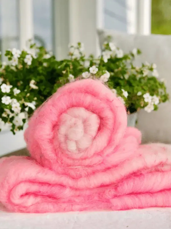 A soft, fluffy pink WoolWomen Art Batt - Corriedale is rolled up on a white surface, with a potted plant of small white flowers in the background—perfect for käsinvärjätty villakuitu and kehrättävä kuitu projects.