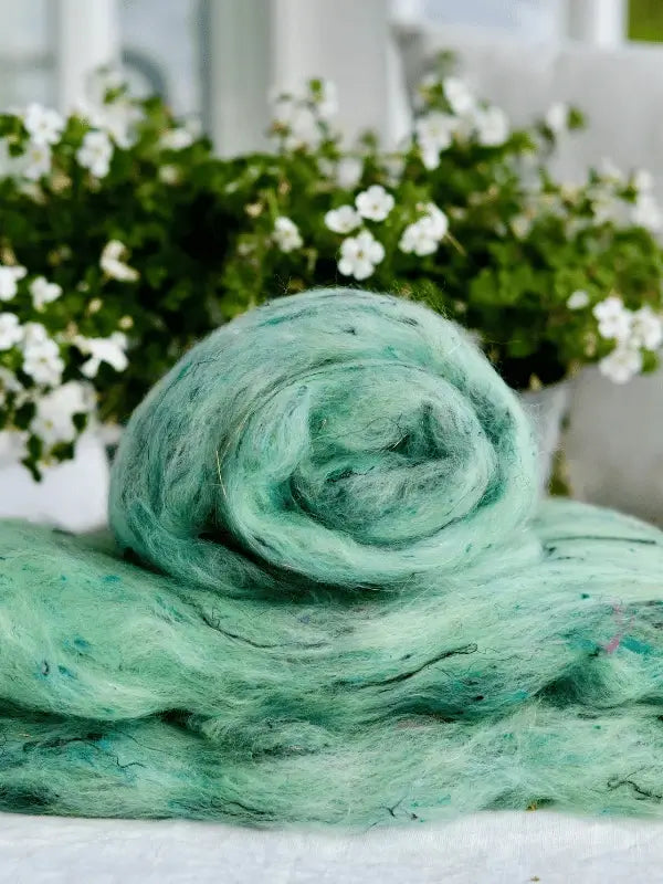 A roll of WoolWomen Art Batt - Corriedale, in soft light green, is displayed on a table with white flowers and green leaves blurred in the background.