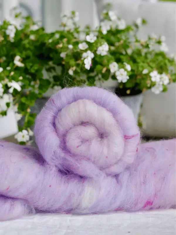 A roll of soft, light purple WoolWomen Art Batt -Corriedale rests on a surface, with white flowering plants blurred in the background.