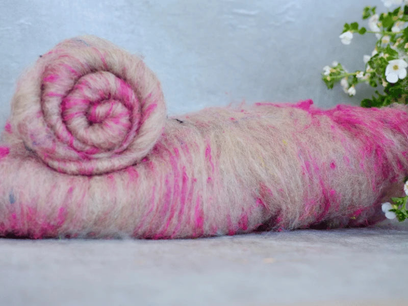 A rolled bundle of WoolWomen Art Batt - BFL with pink streaks rests on a flat surface beside small white flowers and green leaves, set against a light gray background.