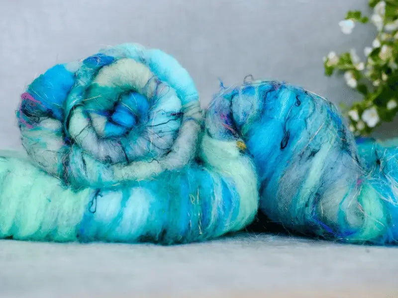 Two rolled bundles of WoolWomen Art Batt -Cormo, featuring streaks of blue and turquoise, lie on a gray surface. A blurred green plant with white flowers appears in the background.