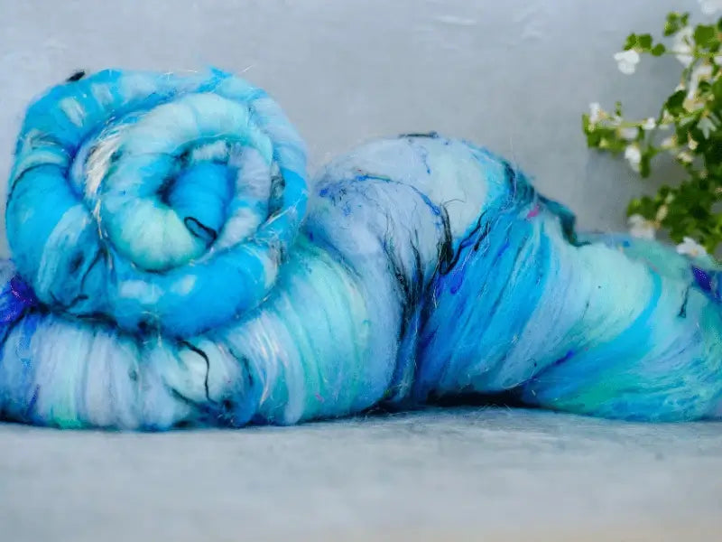 A close-up of two fluffy, rolled WoolWomen Art Batt -Cormo bundles in blue and white with touches of green and purple on a light surface. Small white flowers appear to the right in the background.