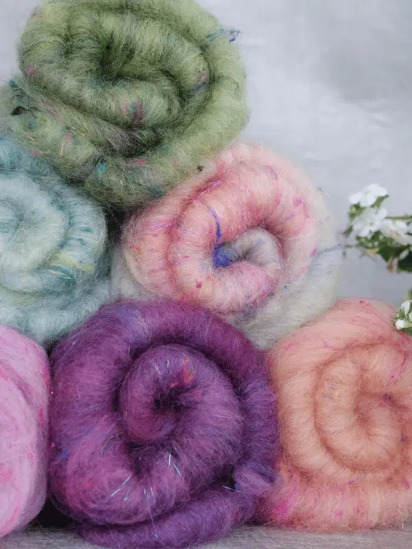Six colorful WoolWomen Art Batt -Suomalainen villa rolls are stacked together, displaying greens, blues, pinks, purples, and peach. A sprig of small white flowers is on the right side against a gray background.