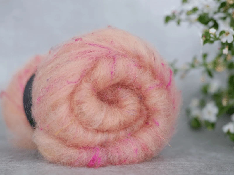 A close-up of a rolled ball of WoolWomen Art Batt -Suomalainen villa in peach-pink shades with brighter pink touches on a light surface, soft white flowers blurred in the background—a great pick for art batt kehräys.