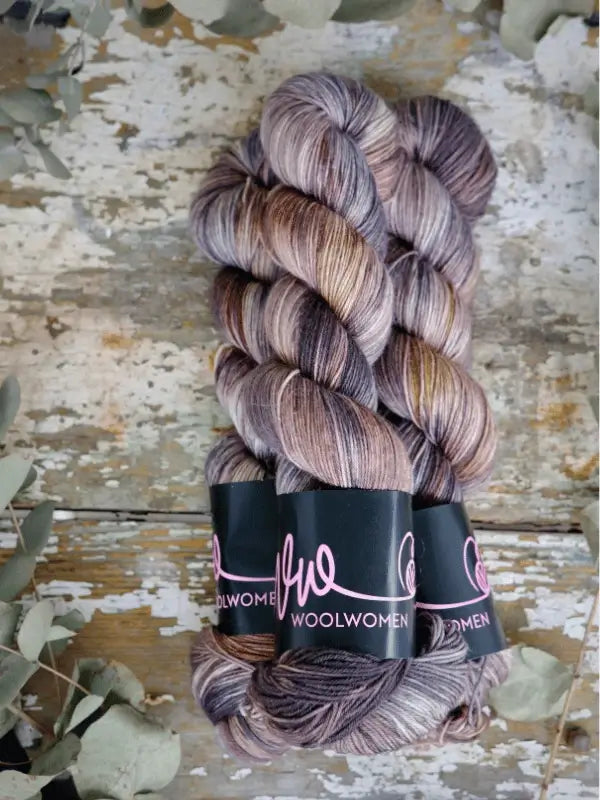 Three skeins of käsinvärjätty lanka in brown, gray, and taupe—each labeled WoolWomen Merino Sock—are arranged side by side on a weathered wooden surface with dried leaves nearby.