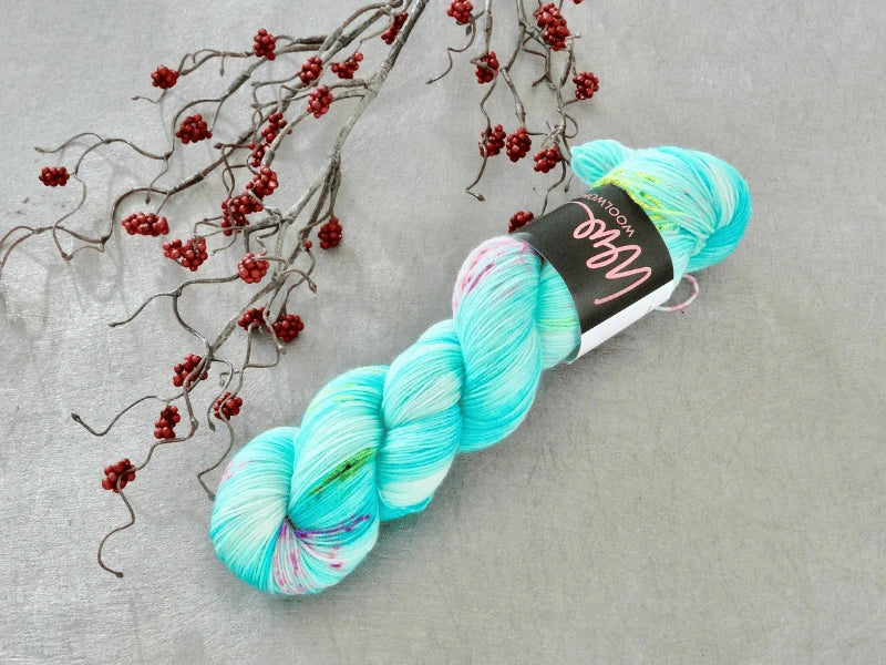 A skein of WoolWomen Merino Sock – Yhden Padan Ihme yarn in turquoise and multicolored hues lies on a grey surface next to a branch with small red berries. This mulesing-free merino sock yarn brings uniqueness to your knitting projects.