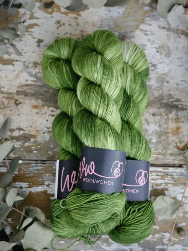 Two skeins of shiny, variegated green WoolWomen Merino Sock yarn rest on a rustic wooden surface, surrounded by dried eucalyptus leaves.