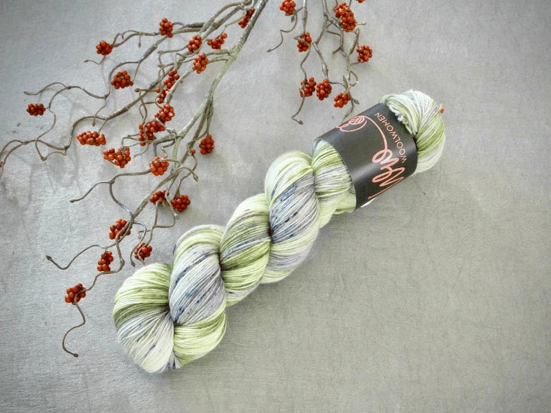 A skein of WoolWomen Merino Sock – Yhden Padan Ihme yarn in light green, white, and gray rests on a gray textured surface beside branches with small orange-red berries.