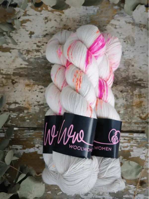 Two skeins of WoolWomen Merino Sock, white with pink and orange speckles, are wrapped in WoolWomen labels and set on a rustic wooden surface with dried leaves—ideal for your next superwash merinovilla project.
