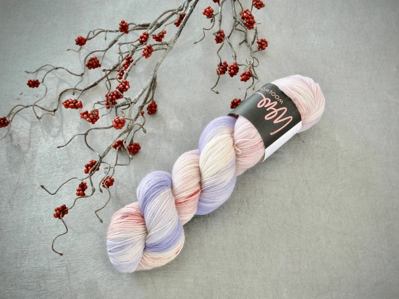 A skein of WoolWomen Merino Sock – Yhden Padan Ihme in pastel pink, lavender, and white lies on a gray surface next to a decorative branch with small red berries.